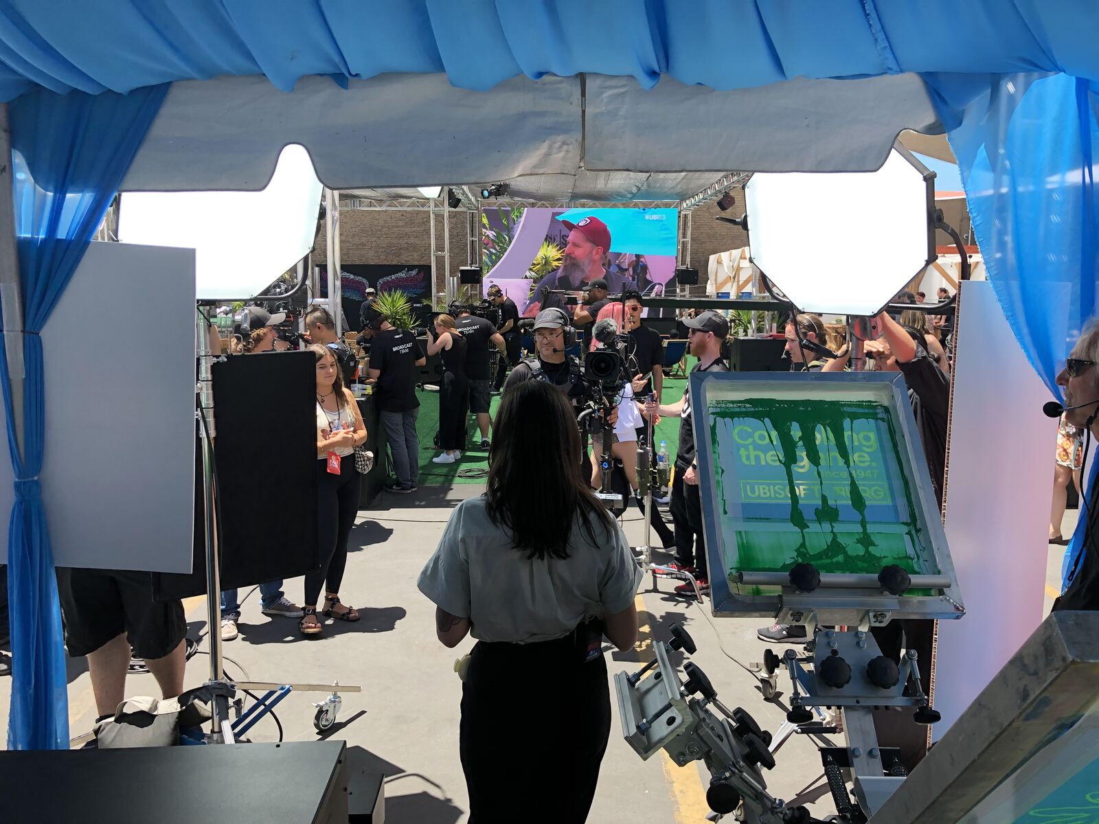 Crowded festival event tent with attendees mingling under a blue canopy with stage and digital displays in the background